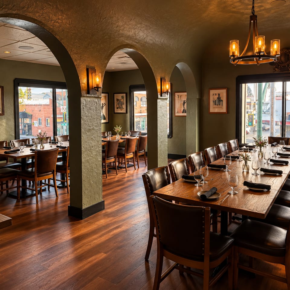 Warmly lit restaurant dining room with arched walls, wood tables, leather chairs, framed artwork, and street views through windows.