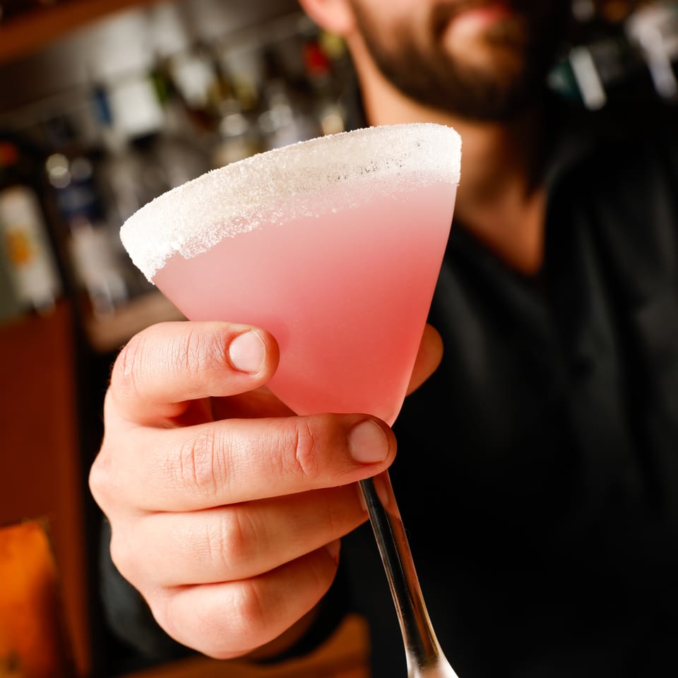 Bartender holds a pink cocktail in a sugar-rimmed glass with a straw, set against a warmly lit bar.