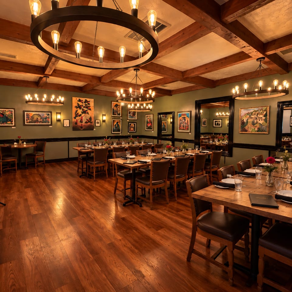 Warmly lit restaurant dining room with wood-beamed ceilings, artistic wall decor, set tables, leather chairs, and circular chandeliers.